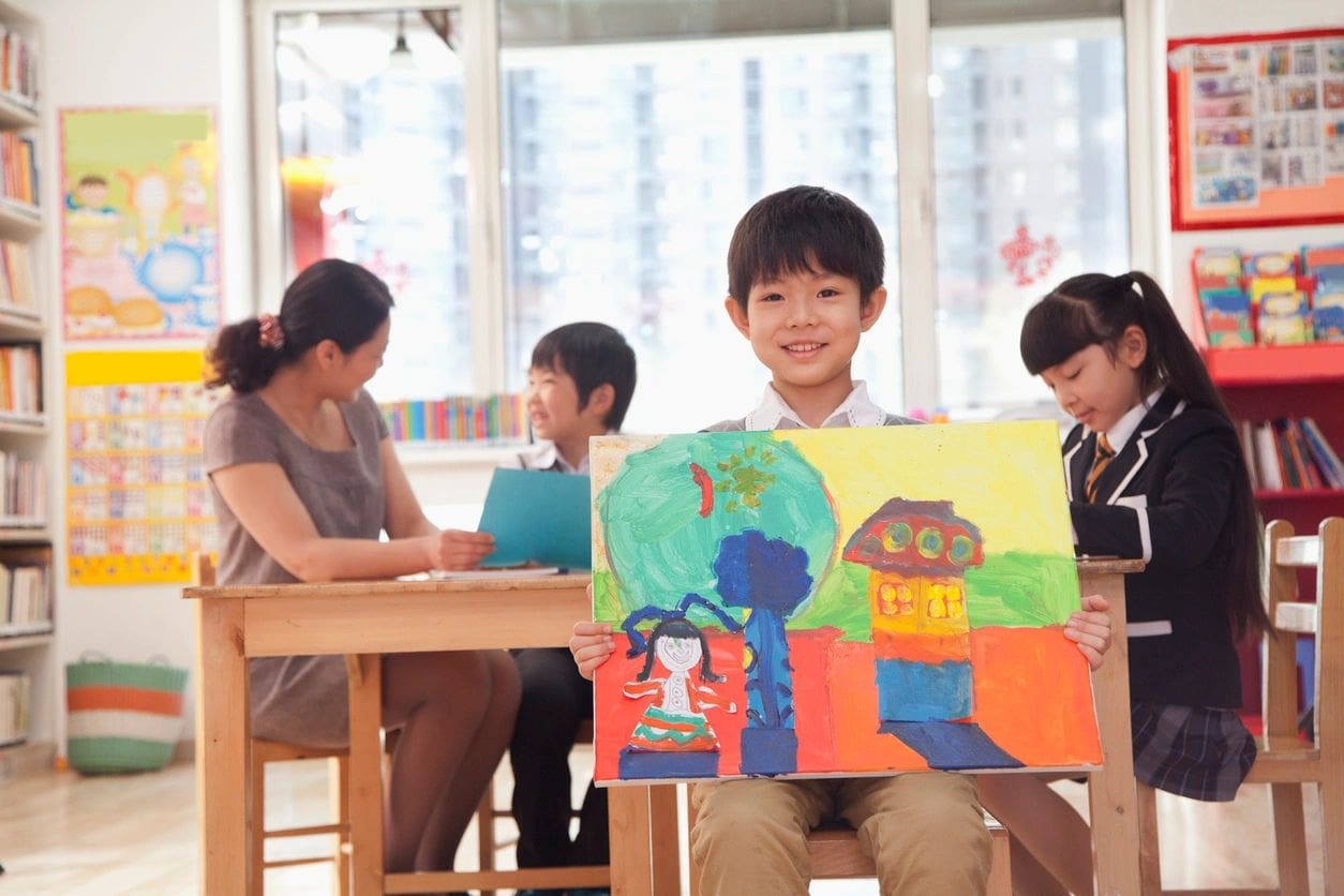 A boy holding up a painting in front of other children.